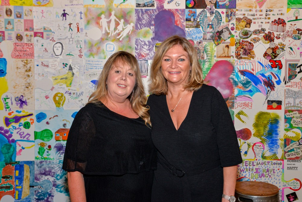 Two women in black posing in front of colourful background