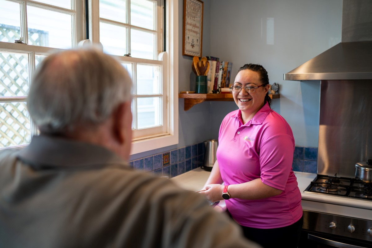 Woman wearing bright pink Feros Care T-shirt laughing at senior man in a kitchen