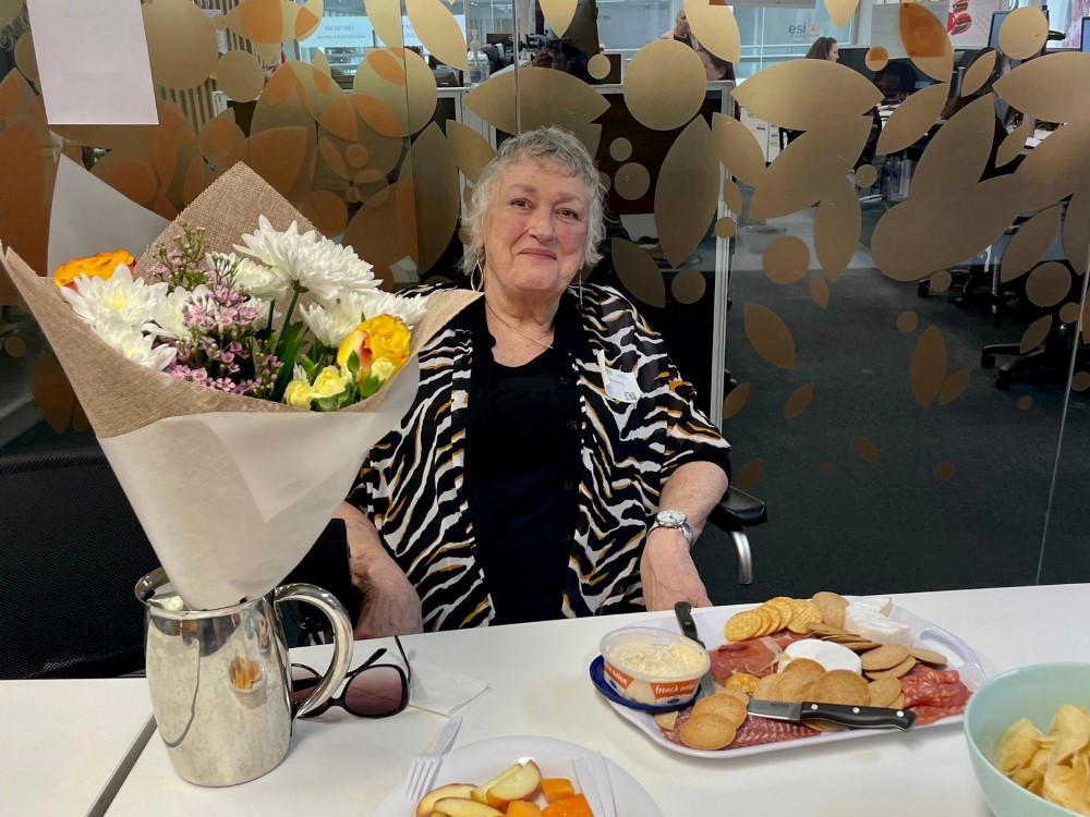 Senior lady sitting at office desk with plate of cheese and crackers and a bouquet of flowers on the table