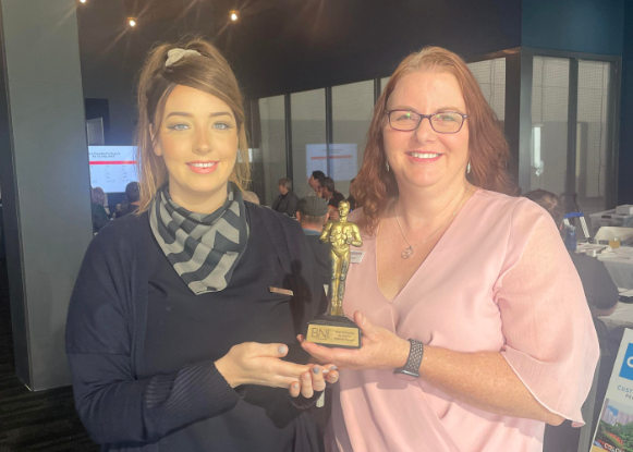 A young woman in black and a middle aged woman in pink holding an award trophy