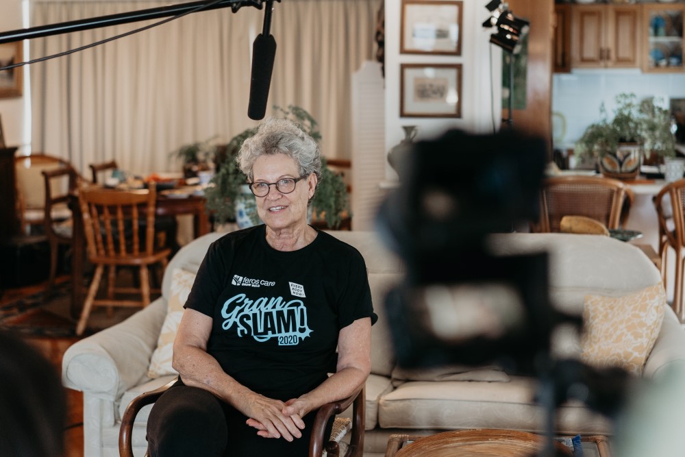 Senior lady wearing black and blue Gran Slam T-shirt and glasses sitting in a chair with a camera in the forefront and a mic above her