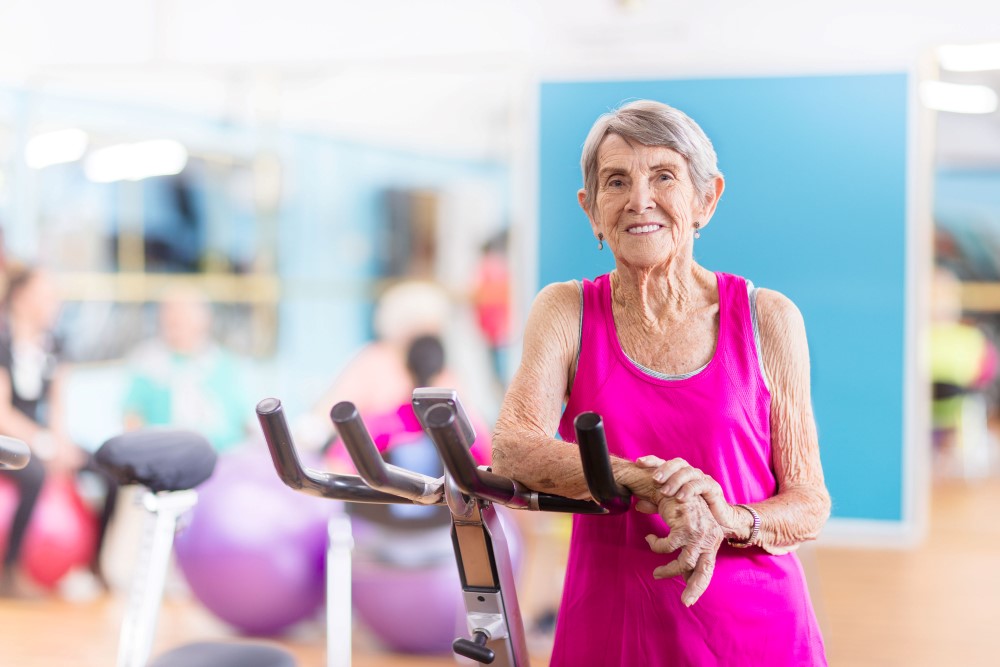 Smiling grandma wearing pink while standing beside stationary bike 