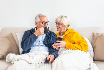 An elderly couple cozy on a couch under a blanket, with mugs in hand