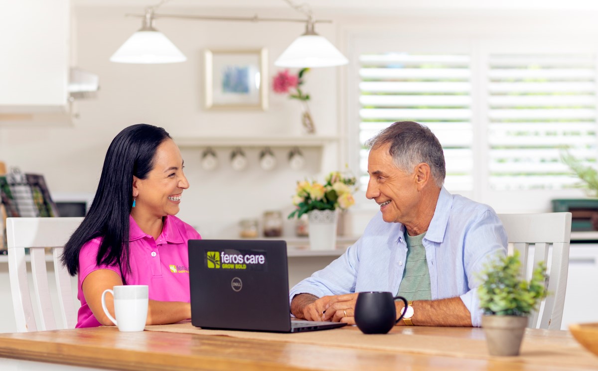 Young woman with black hair and pink T-shirt and senior man in blue shirt smiling at each other at a table in front of a Feros Care laptop