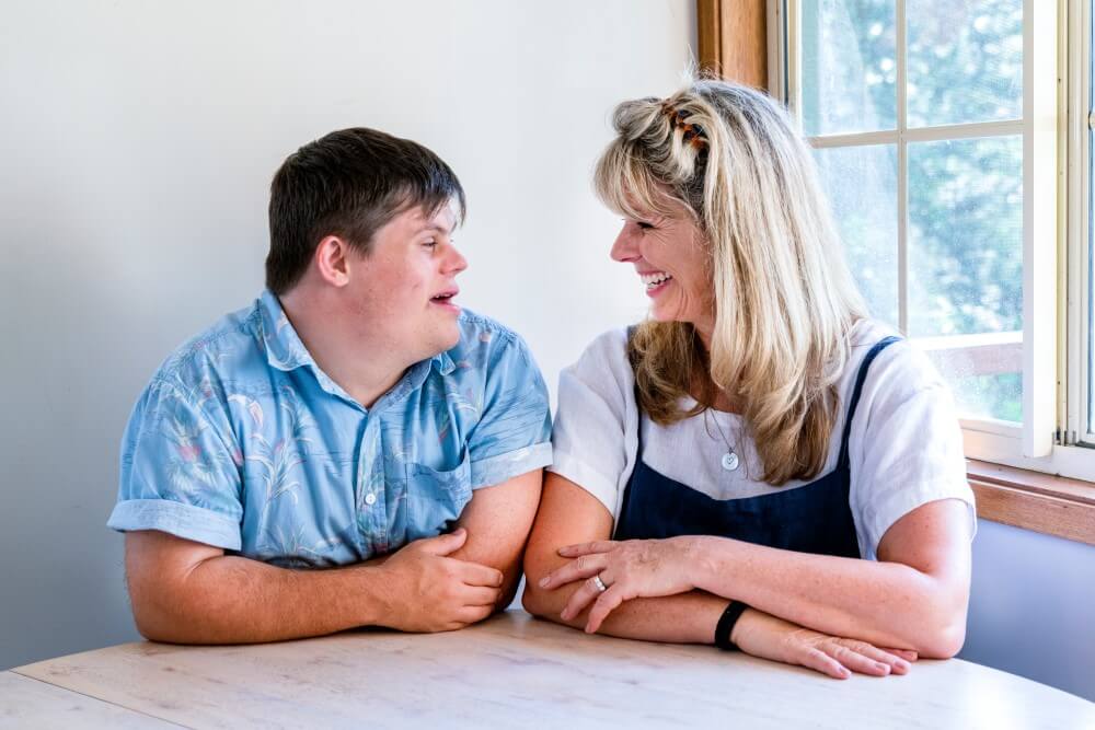 Teenager with Down syndrome and his mum smiling at each other at a table