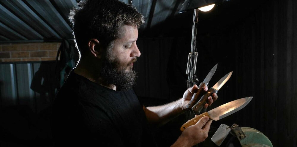 Man in a dark shed holding the knives he's made