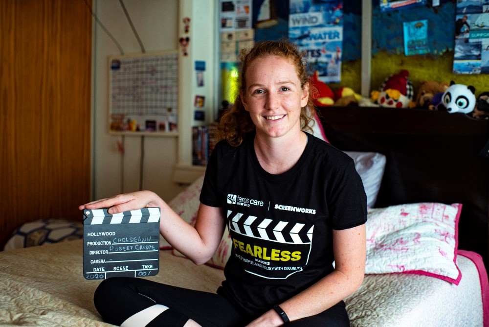 Girl with red hair sitting on her bed in a black Fearless T-shirt, holding a clapboard