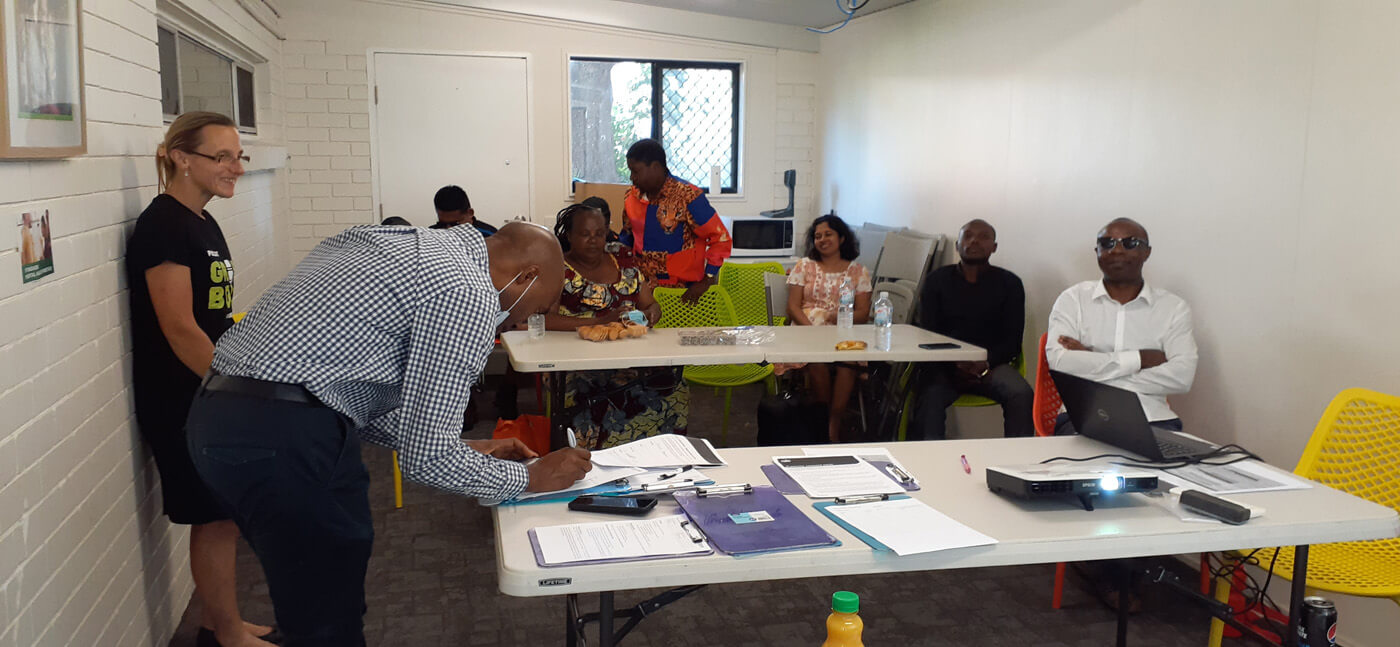 8 people in a room. Most people are sitting looking toward the camera. One man is bent over completing paperwork.
