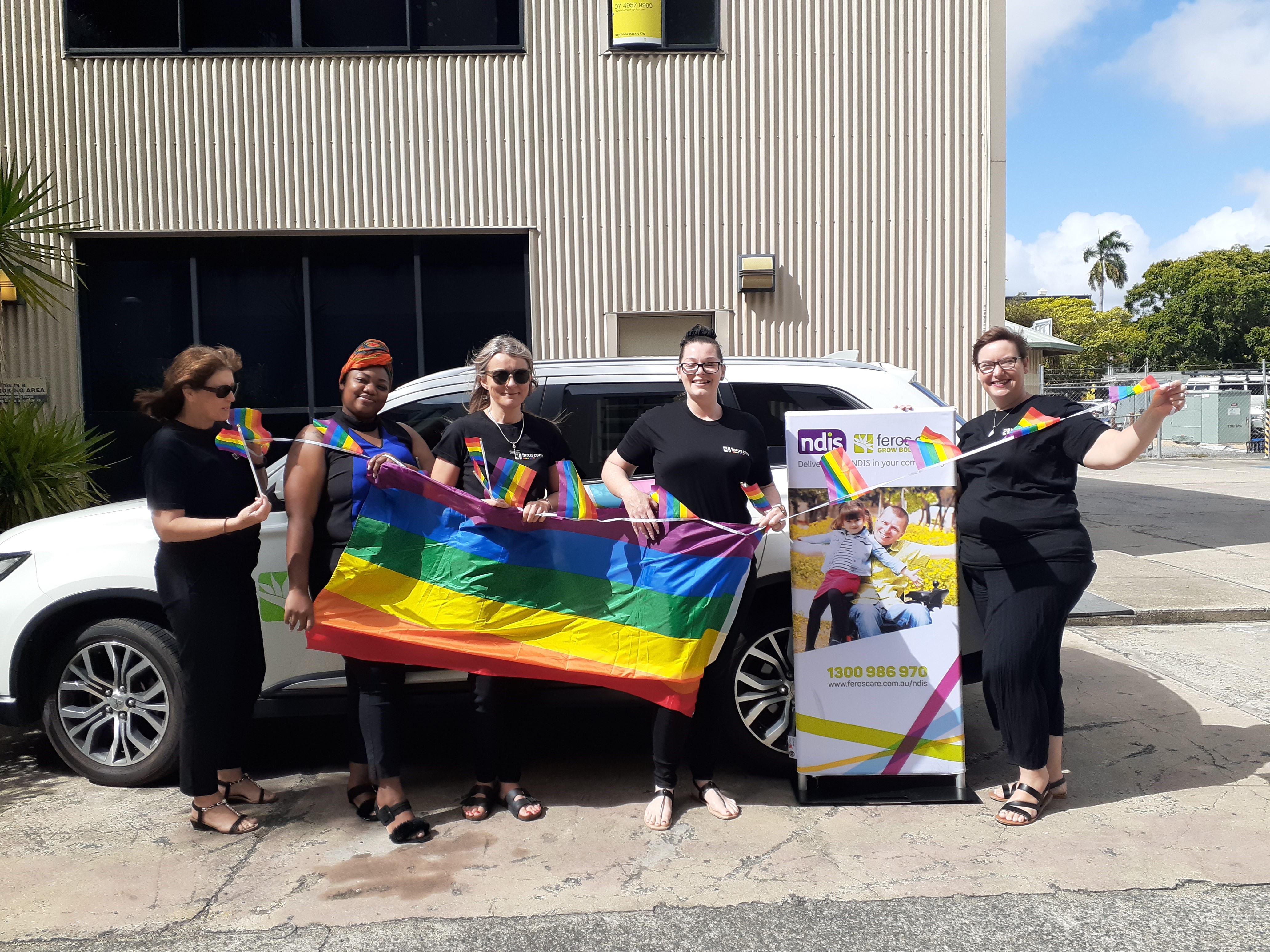 Five women standing in front of car holding a big pride flag