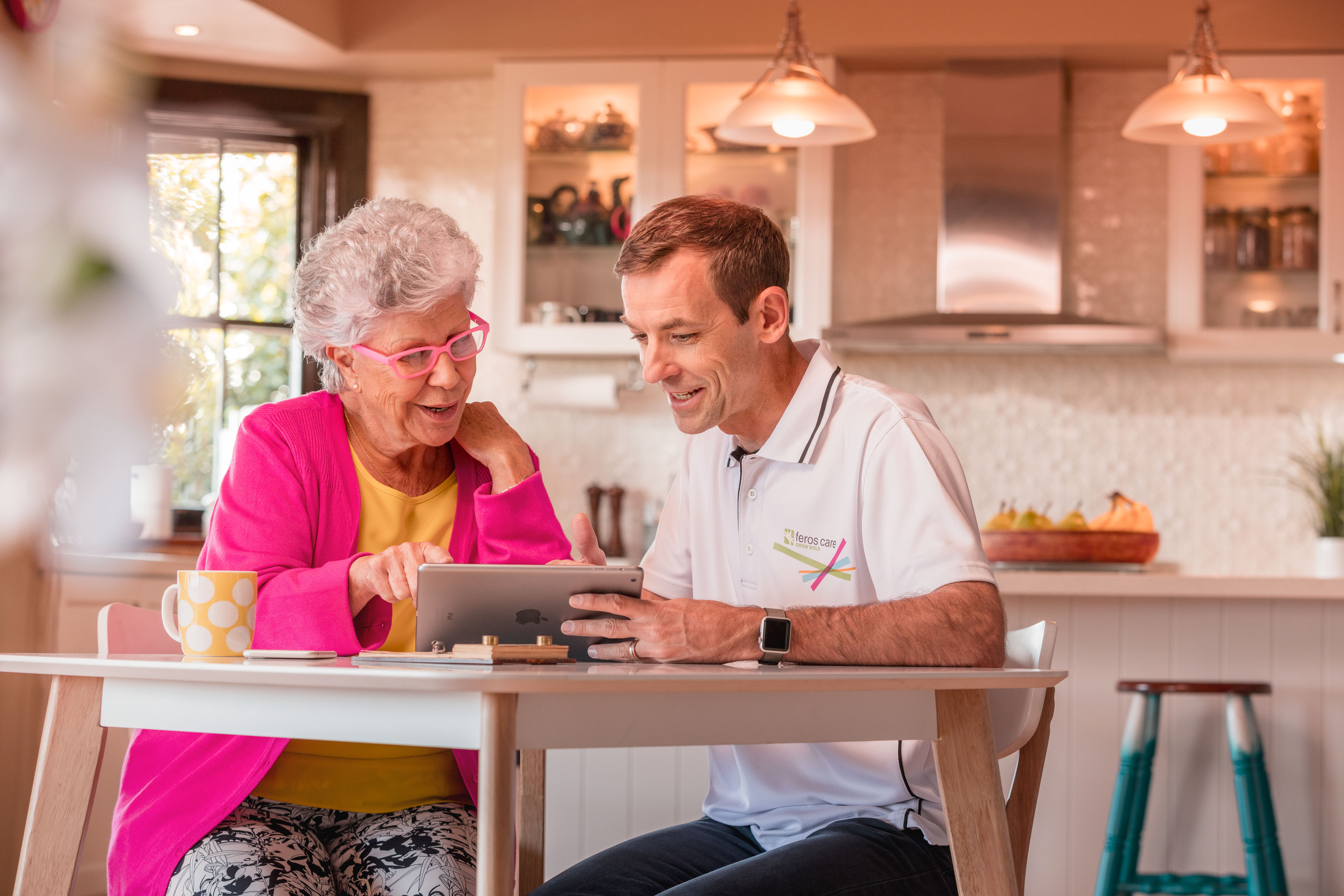 Lady with pink cardi on tablet with Feros Care man