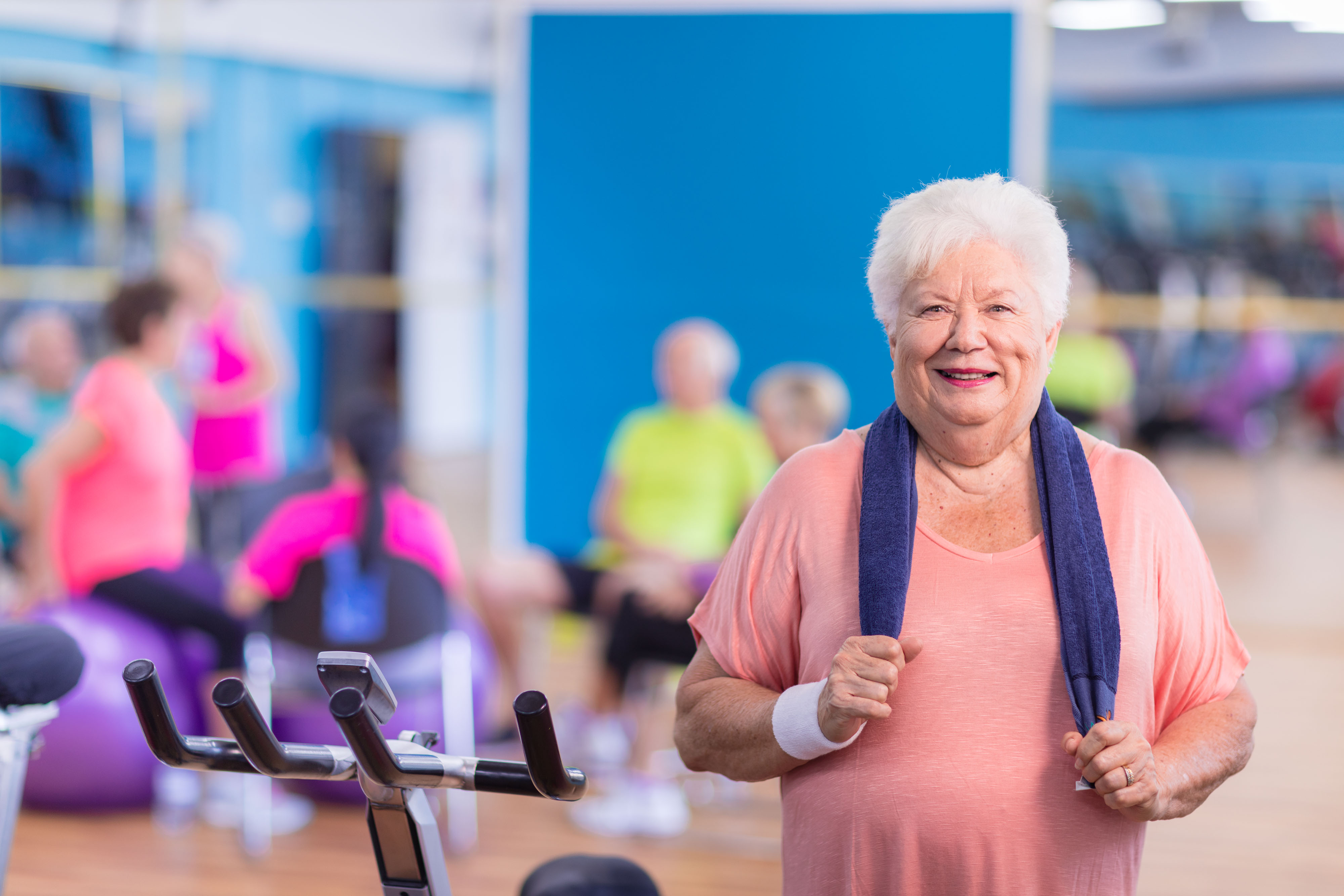 Grandma smiling wearing peach shirt holding blue towel - standing beside stationary bike