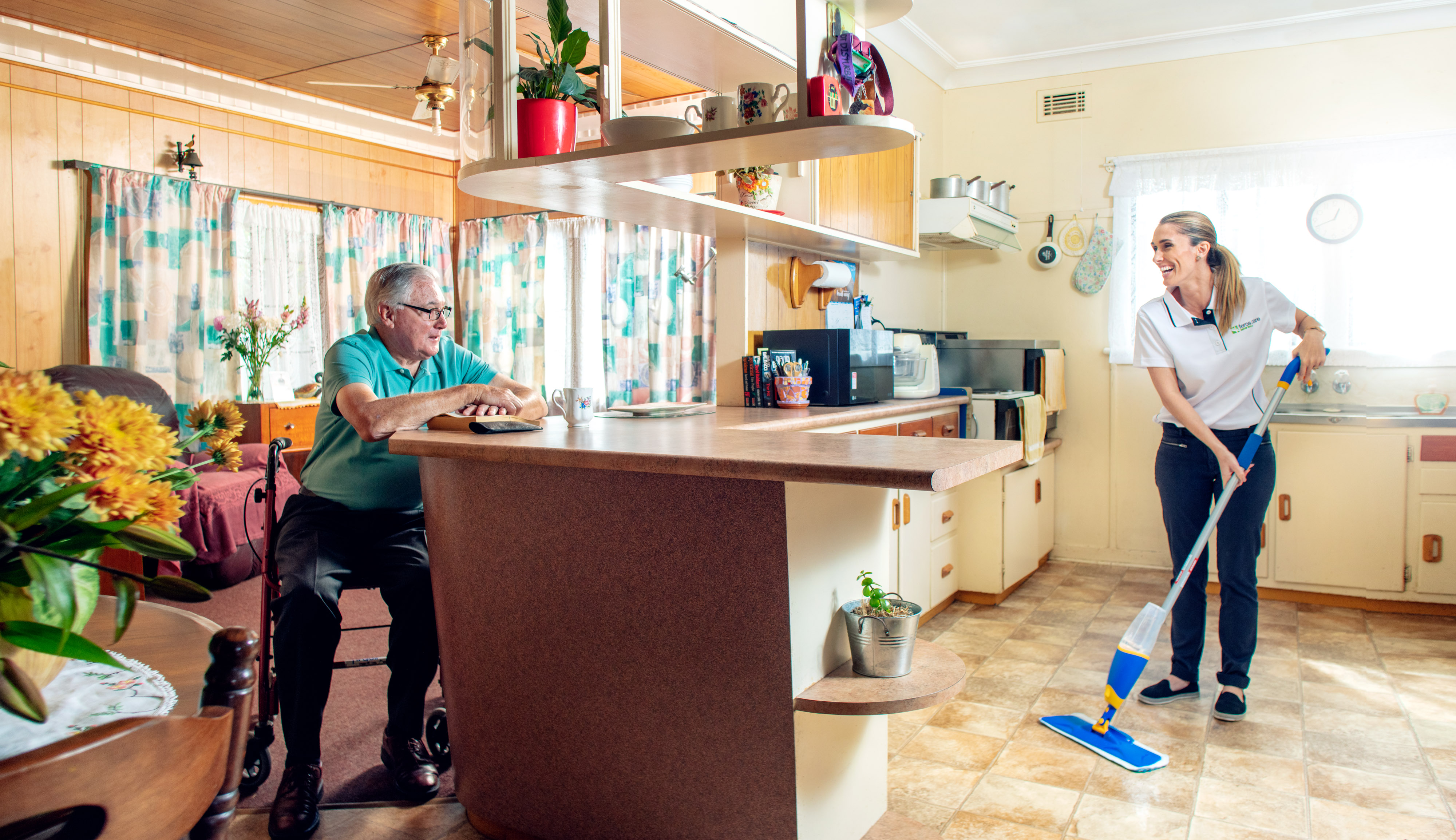 Senior man sitting on bar stool while care worker cleans kitchen