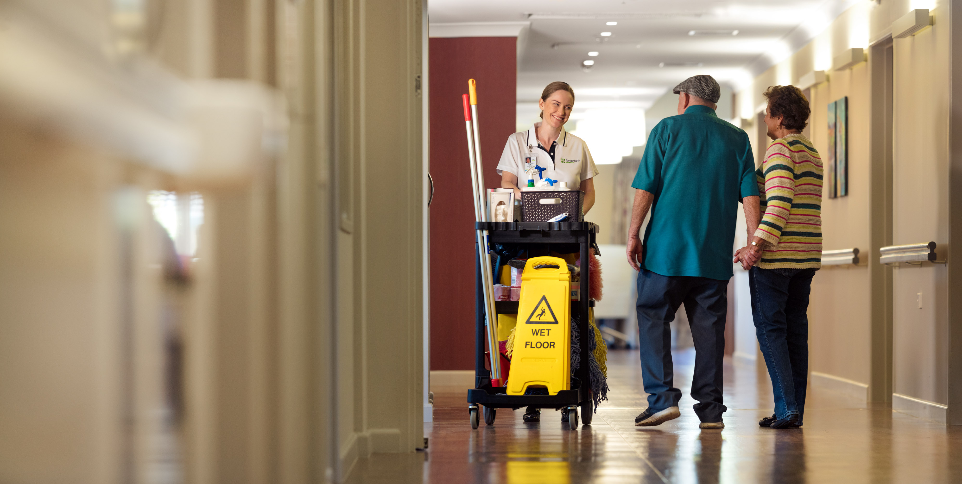 Cleaner passing two seniors in aged care home corridor