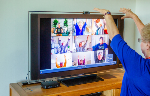 Elderly lady sitting in front of TV and doing stretches with others virtually