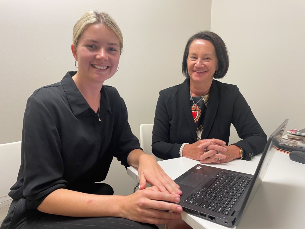 Young woman with blonde hair in a pony tail and middle aged woman with black bob seated at a desk with a laptop in front of them