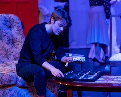 Young queer person in a blue-tinted room seated on a couch and looking at chess set on a coffee table