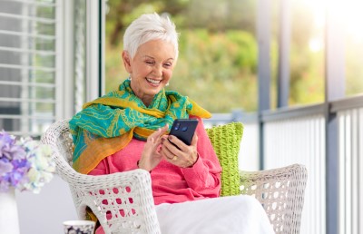Senior lady with short white hair wearing pink top and colourful scarf sitting on a patio chair and smiling at her phone