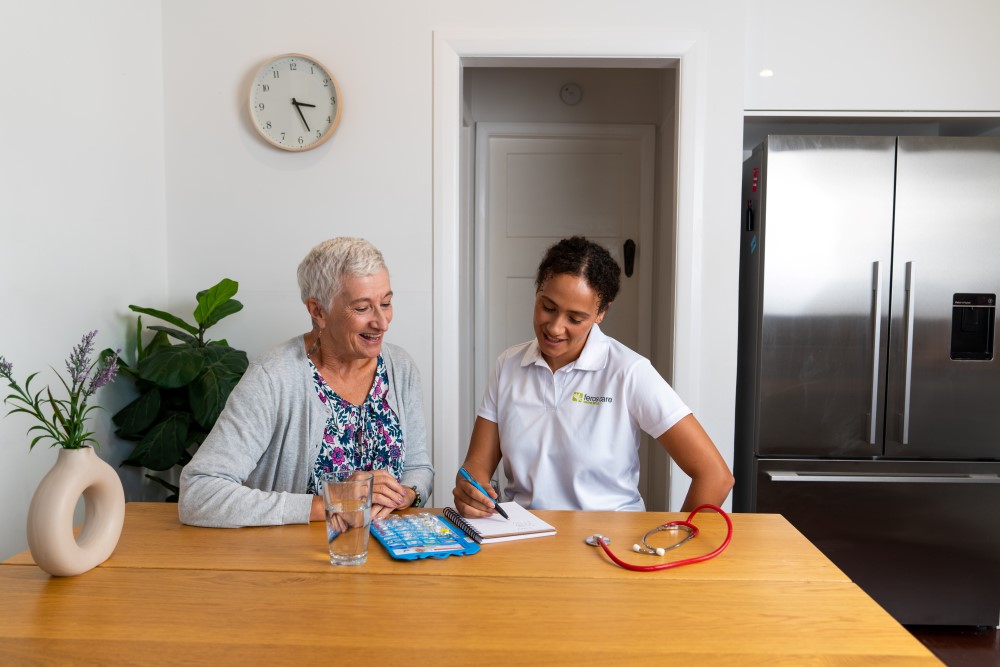 Senior lady and young healthcare professional seated at kitchen table, looking through notes and medications