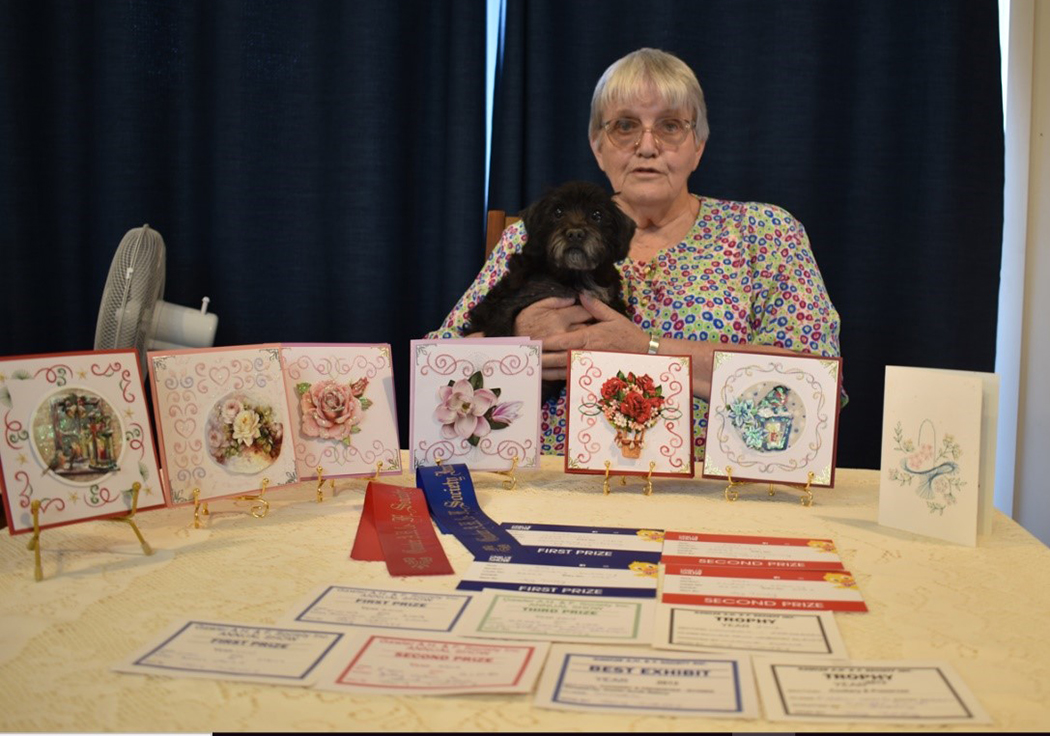 Joy and her pet dog sitting behind a table with her handmade cards and Gawler show first and second prize ribbons and certificates..