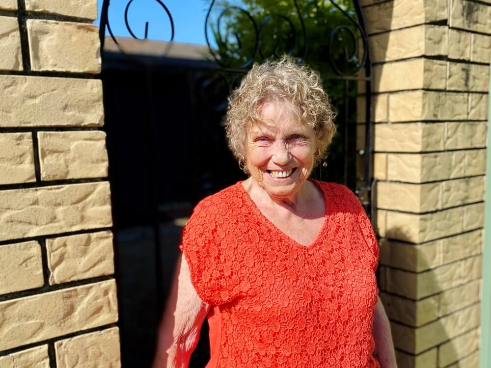 Senior lady in bright orange top smiling in front of a gate