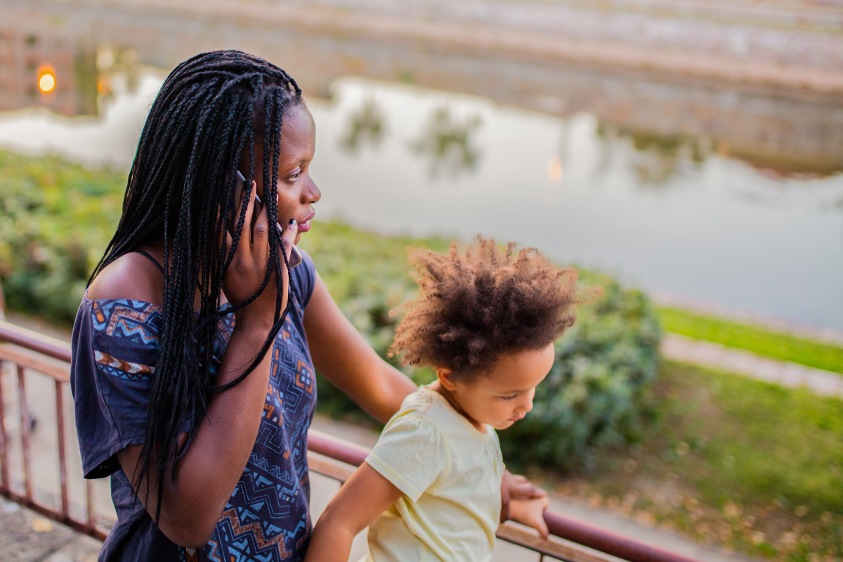 A Black woman with box braids on the phone, looking worried, with a young child in front of her