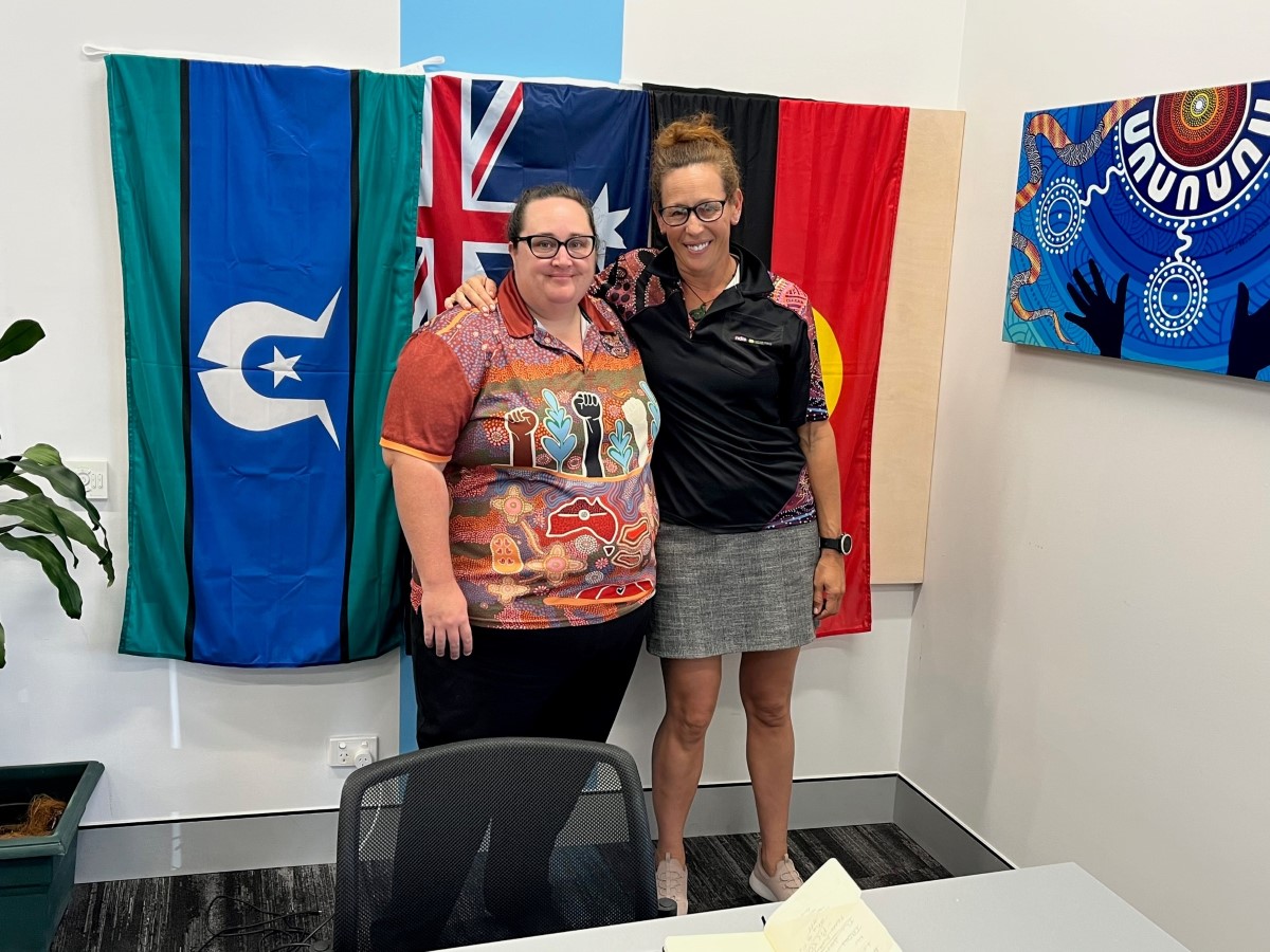 Two women wearing glasses and polo shirts with Aboriginal art on them posing for a photo in front of the Torres Strait Islander, Australian, and Aboriginal flags. 