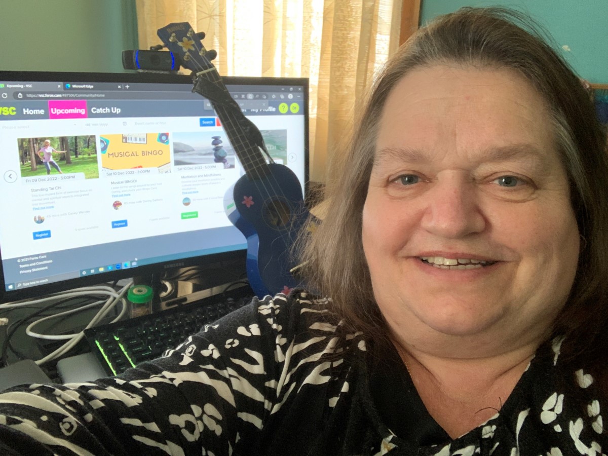 Middle-aged lady taking a selfie in front of a computer screen, with a ukulele leaning against it 