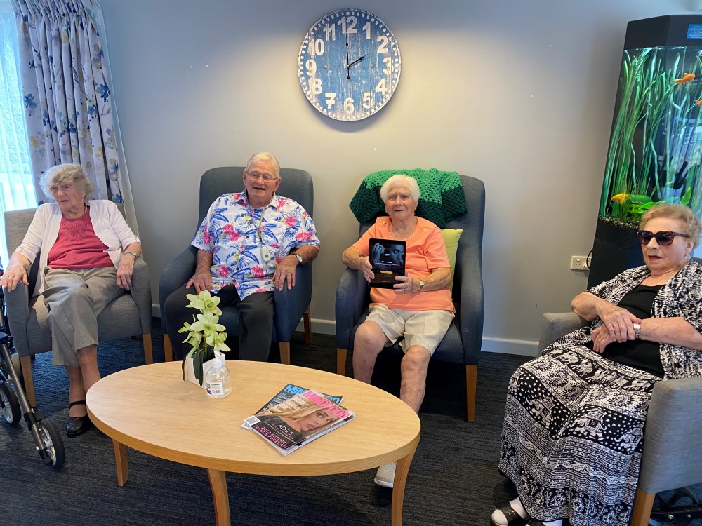 Four seniors - one man and three women - seated in armchairs, one of the ladies holding up a tablet