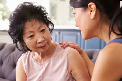 Senior Asian lady and young Asian woman sitting on couch, with the woman's hand on the lady's shoulder