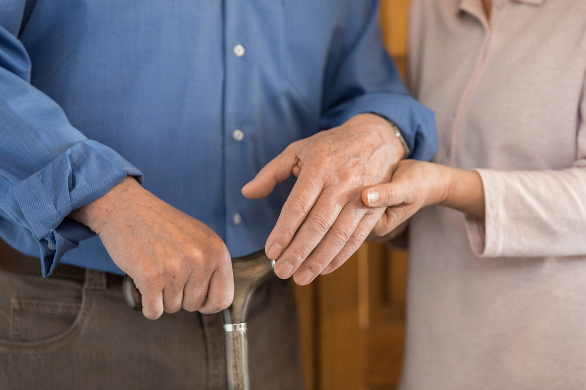 A younger woman's hands holding a senior man's hands as he holds onto a walking stick