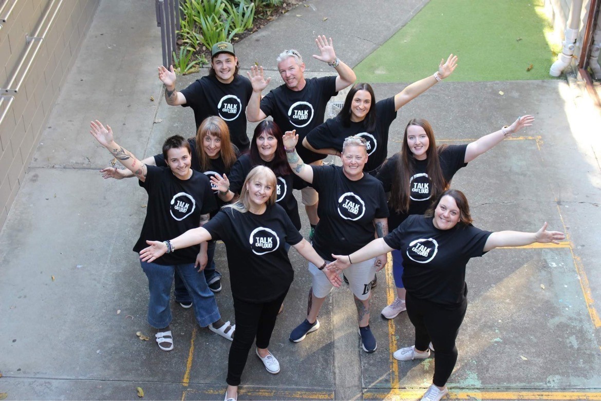 A group of people all in black Talk Out Loud T-shirts with their arms up posing for a photo