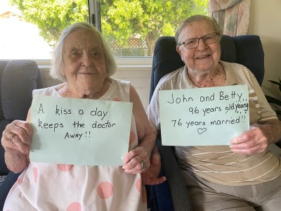 Senior couple sitting next to each other, holding signs. One says "A kiss a day keeps the doctor away!" and the other says "John and Betty, 96 years young, 76 years married!"